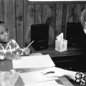 Black and white photo of teacher sitting at table with student in classroom almost 35 years ago
